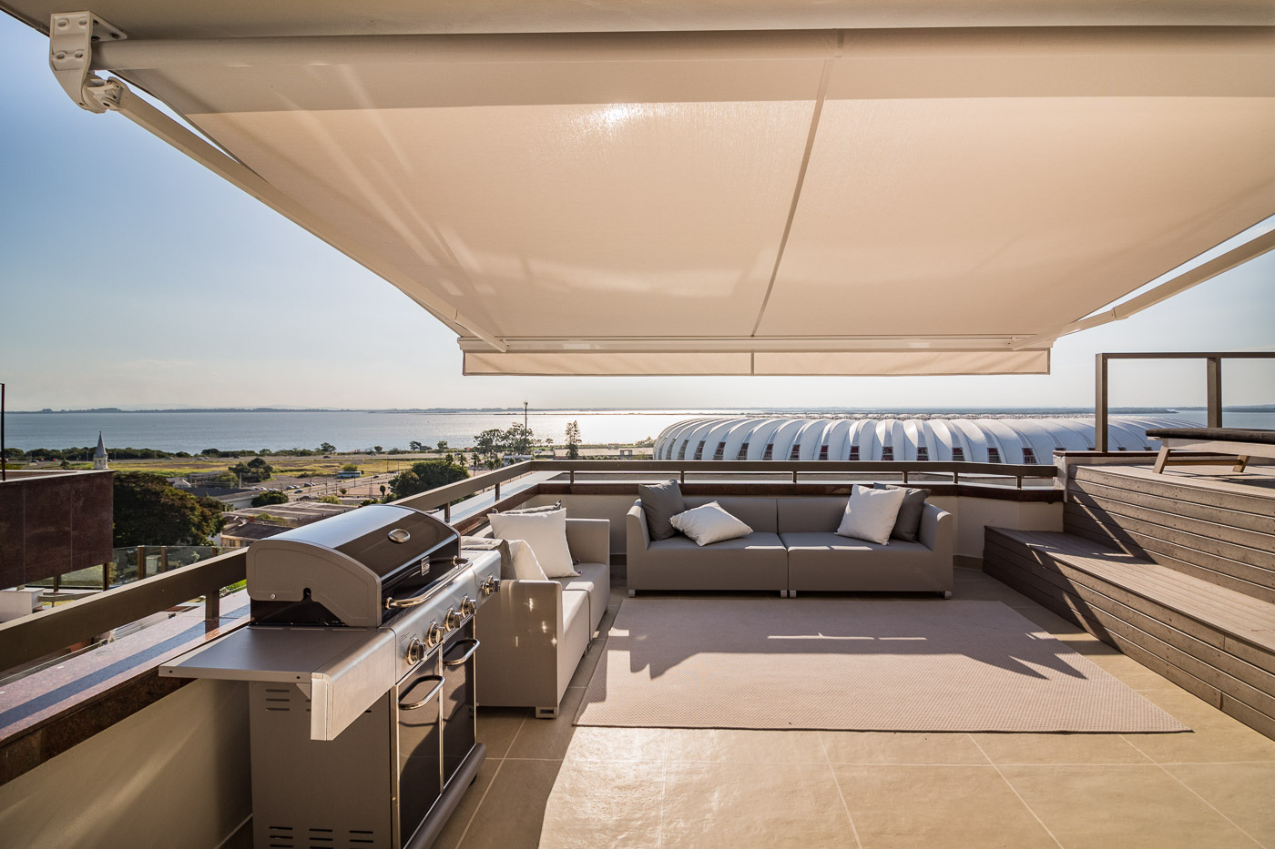 Terraço com vista panorâmica para o mar e estádio, toldo retrátil bege articulado protegendo área de lazer com sofás cinza e churrasqueira a gás, luz dourada de fim de tarde, fotografia arquitetônica profissional