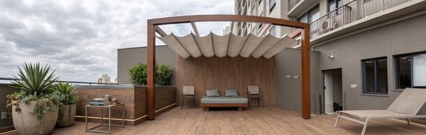 Pérgola retrátil de madeira em terraço de cobertura, lona bege articulada, ambiente lounge com sofá cinza e plantas em vasos, piso de deck de madeira, vista de prédios ao fundo, fotografia arquitetônica realista