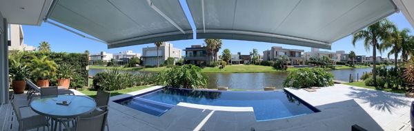 Toldo articulado branco panorâmico instalado sobre área externa de casa com piscina, vista para lago e casas modernas ao fundo, palmeiras e jardim tropical, dia ensolarado, fotografia arquitetônica realista