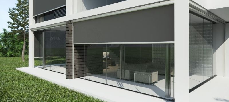 Toldos motorizados retráteis em tom bege instalados lado a lado sobre fachada de casa escura, área externa com mesa e cadeiras estofadas, aquecedor de ambiente, fotografia realista noturna