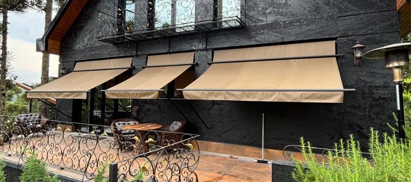 Toldo de braços retos com lona bege instalado sobre terraço com vista panorâmica de bairro residencial, sofás externos cinza, dia ensolarado, fotografia realista