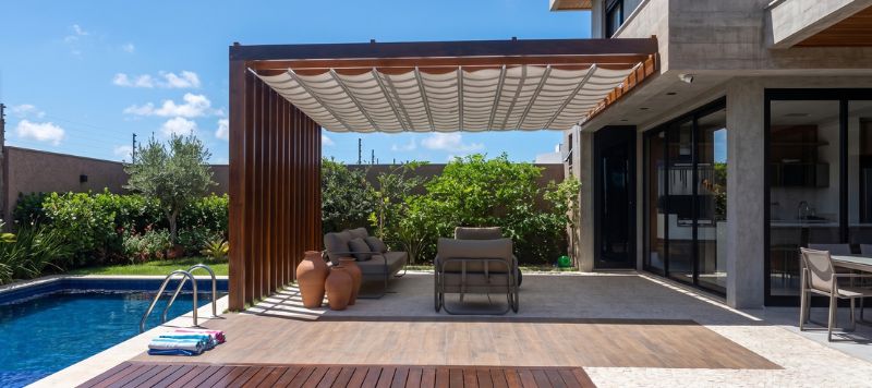 Toldo pérgola retrátil de madeira instalado junto à piscina, tecido dobrável bege, ambiente externo sofisticado com sofás e vasos decorativos, dia ensolarado, fotografia arquitetônica realista