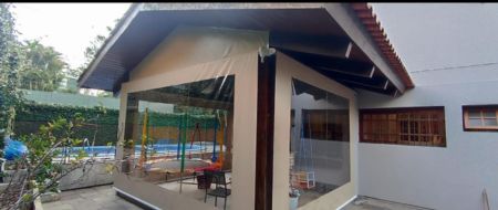 Área de lazer coberta com toldo articulado bege sobre piscina, estrutura de madeira e vidro, jardim com plantas ao redor, dia nublado, fotografia arquitetônica realista