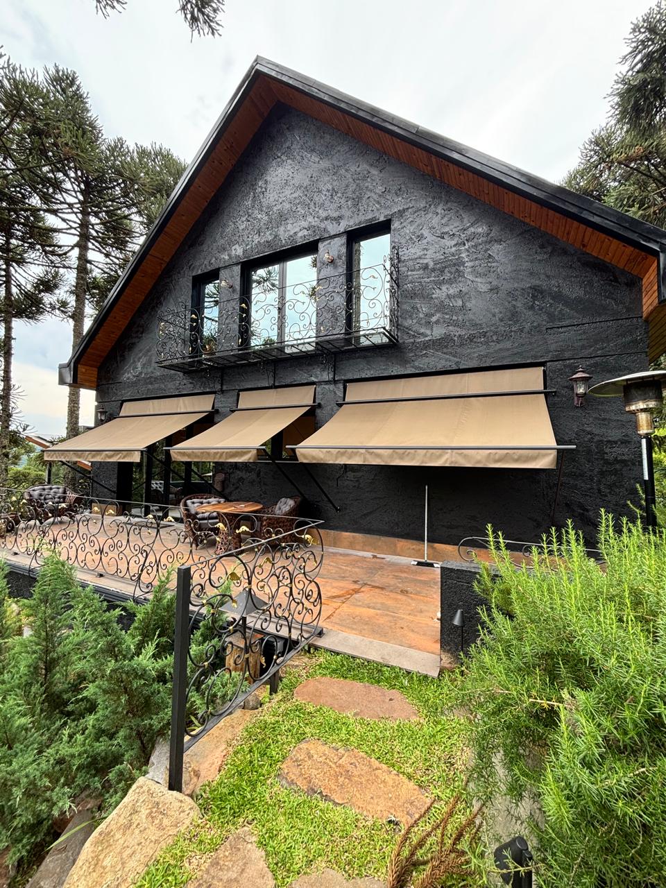 Casa estilo chalé com fachada preta e detalhes de madeira, três toldos articulados bege instalados sobre deck com móveis de área externa, cercada por pinheiros e jardim verde exuberante, luz natural diurna, fotografia arquitetônica realista