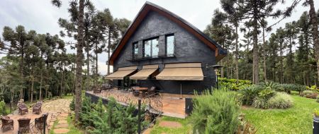 Casa moderna estilo chalé com fachada preta e detalhes em madeira, toldo articulado bege instalado sobre deck de madeira, cercada por pinheiros e jardim verde exuberante, área externa com mesa e cadeiras, fotografia realista de arquitetura