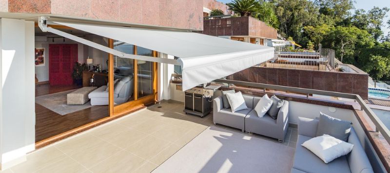 Toldo retrátil articulado instalado sobre varanda residencial, estrutura de madeira e lona translúcida, área externa com sofás e mesa de jantar, jardim ao fundo, fotografia realista