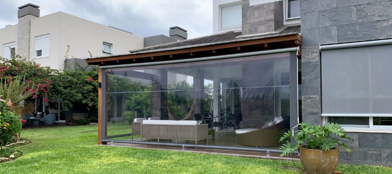 Toldo vertical retrátil cinza instalado em fachada moderna com vidro, fechando lateral de área externa, paleta neutra, fotografia arquitetônica realista
